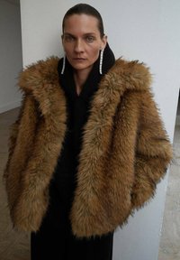 Cappotto in finta pelliccia marrone con un ampio cappuccio, caratterizzato da una superficie testurizzata e un design morbido, indossato su un indumento nero, abbinato a orecchini lunghi.