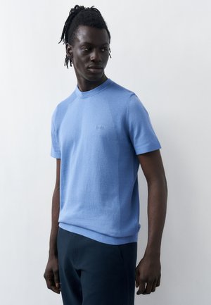 Άνδρας με dreadlocks που φοράει ένα ανοιχτό μπλε πλεκτό κοντομάνικο πουκάμισο και σκούρο παντελόνι, στέκεται μπροστά σε απλό ανοιχτό φόντο.