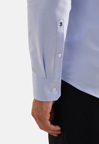Camicia azzurra con una texture liscia, caratterizzata da polsini a punta con bottoni bianchi e dettagli ricamati di rose nere.