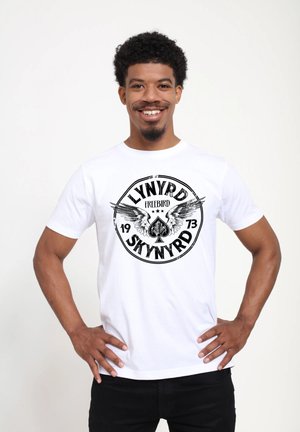 T-shirt en coton blanc avec un graphique noir circulaire portant le texte "LYNYRD SKYNYRD FREEBIRD 1973" et des motifs ailés. Texture lisse.