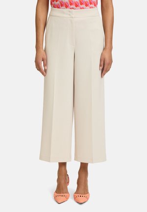 Wijdvallende broek van lichtbeige stof, met een hoge taille en scherpe plooien. Gecombineerd met textuur, koraalkleurige schoenen.