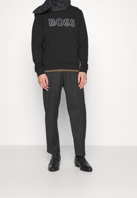 Černá mikina s bílým logem „BOSS“, doplněná šedými pánskými kalhotami a černými kotníkovými botami. Okolo krku je přehazena černá šála.