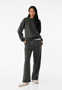 Kuldgrå sweatshirt og matchende vide bukser. Blødt stof med en afslappet pasform. Hvid t-shirt lag synlig under sweatshirten.