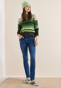 Pull en maille rayé vert et beige, jean foncé et bonnet vert. Contrastes de textures dans les matériaux, avec des motifs rayés et des baskets décontractées.