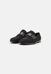 Zapatos negros de tipo slip-on con parte superior de tela texturizada y correa de acento negra brillante. Diseño de punta afilada y tacón bajo plano.
