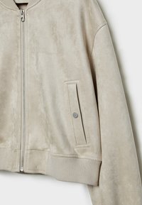 Beige bomberjakke lavet af blødt materiale, der minder om ruskind. Den har en lynlås foran, sidelomme med knap og ribstrikket kant og manchetter.