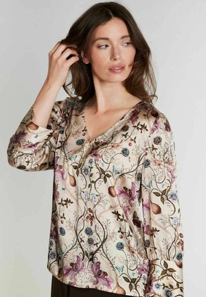 Femme aux longs cheveux bruns portant un chemisier crème avec un motif floral et de papillons, regardant sur le côté tout en touchant ses cheveux.