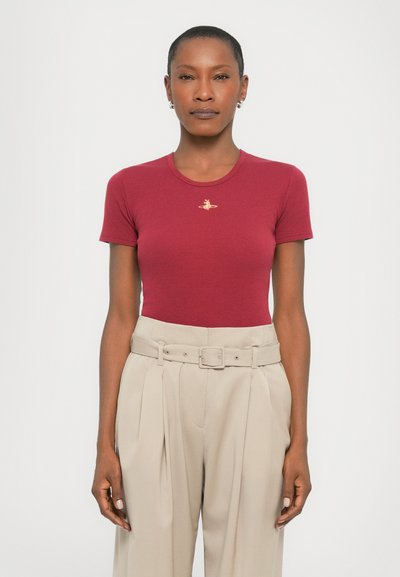 Donna che indossa una maglietta attillata a maniche corte color bordeaux con un piccolo emblema e pantaloni beige a vita alta con pieghe e una cintura abbinata.