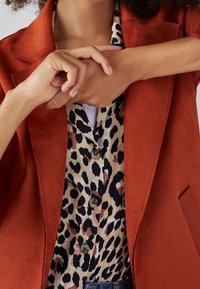 Blazer de color óxido superpuesto a una blusa de estampado de leopardo con acentos en negro y marrón. Textura suave, se ven dos botones en la blusa.