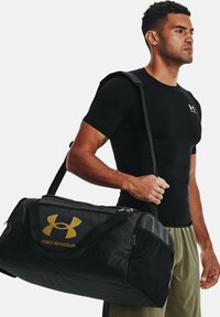Juodas sportinis krepšys su auksiniu Under Armour logotipu, turintis užtraukiamą viršų, tvirtas rankenas ir tekstūruotą paviršių. Neša vyriškas modelis.