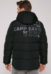 Giacca imbottita nera con cappuccio, caratterizzata da lettering prominente "CAMP DAVID" e coordinate geografiche sul retro. Tessuto testurizzato e dettagli cuciti.