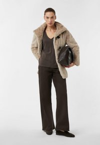 Beżowy płaszcz z sztucznego futra z dużym kołnierzem, ciemnobrązowa bluzka i spodnie o szerokich nogawkach, trzymająca ciemnobrązową torebkę. Miękkie tekstury i neutralne kolory.