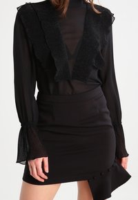 Outfit nero composto da una blusa trasparente con scollo arricciato e maniche lunghe texturizzate, abbinato a una minigonna aderente con dettagli ai bottoni.