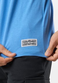 Blå t-shirt med kort ärm i mjukt material, med en vit rektangulär etikett med svart text i nederkanten. Svarta byxor syns.