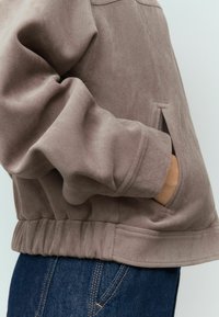 Taupe nepbont leren jas met een geribde zoom, zijzakken en ontspannen mouwen, gecombineerd met donkere spijkerbroeken. Glad textuur.