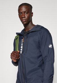 Marineblauwe lichtgewicht jas met een capuchon, voorzien van een ritssluiting aan de voorkant, logo op de mouw en een textuurstof. Het model draagt een groene rugzak.