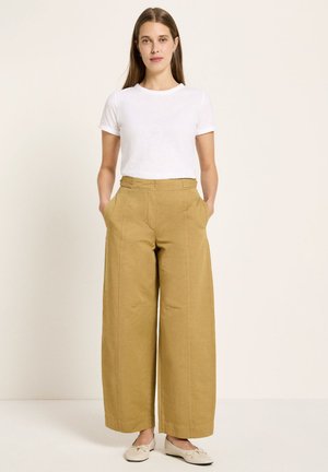 Woman standing with hands in pockets, wearing a white short-sleeve shirt, wide-leg tan pants, and beige flat shoes against a plain background.