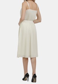 Abito midi plissettato in crema con spalline regolabili, vita aderente e gonna fluente. Tacchi alti neri completano l'outfit.