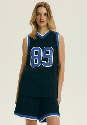 Młoda kobieta o blond włosach, ubrana w czarny bezrękawnik do koszykówki i pasujące spodenki z niebiesko-białą lamówką, z numerem 89 z przodu.