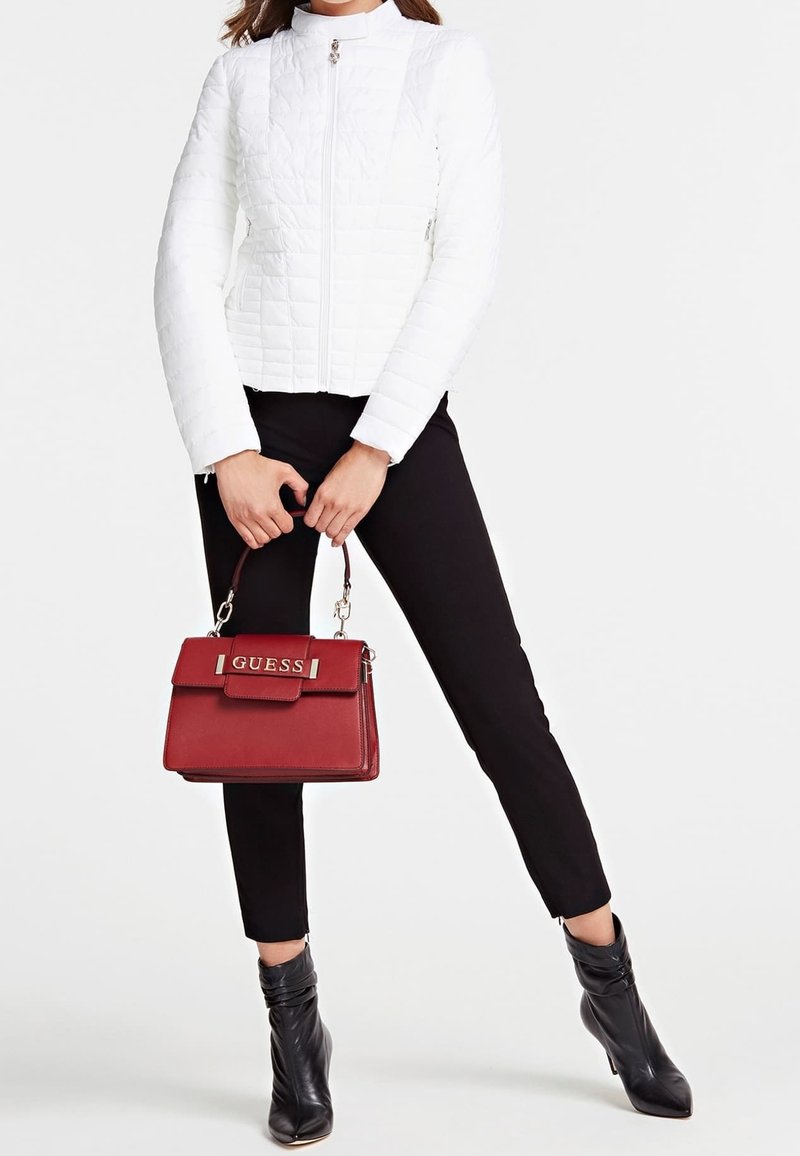 Femme en veste matelassée blanche, pantalon noir et bottines noires tenant un sac à main rouge Guess avec des poignées supérieures.
