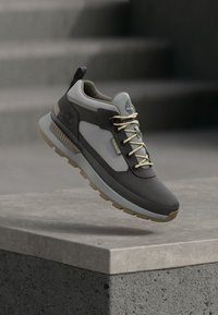 Γκρι και μπεζ αθλητικό παπούτσι με υφασμάτινο πάνω μέρος, επενδυμένο γιακά, αντιθετικές λεπτομέρειες και ελαφριά λαστιχένια σόλα. Τα κορδόνια είναι μπεζ με μεταλλικές άκρες.