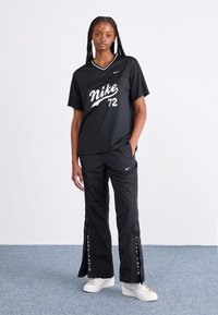 Črna Nike majica s belim grafičnim napisom "Nike 72" in črne hlače z zadrgo in logotipom. Obleka ima sproščen kroj in lahkotno material.