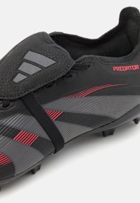 Black soccer cleat with textured synthetic upper, gray and red stripe pattern, elastic band, and distinct three-stripe logo on the tongue.