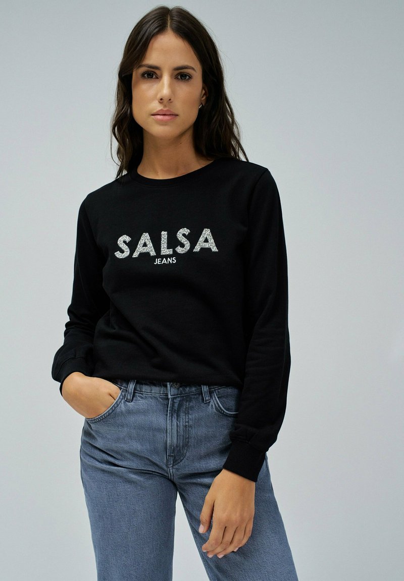 Sudadera negra con el texto "SALSA JEANS" en plata, combinada con unos vaqueros de denim azul claro. La tela parece suave con puños acanalados.