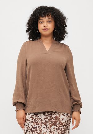 Blouse marron à manches longues avec un décolleté en V et un tissu texturé. Présente des poignets élastiqués et une coupe confortable. Associée à une jupe à motifs.
