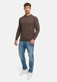 Homme se tenant debout, portant un pull en tricot marron, un jean bleu et des baskets blanches, contre un fond clair.