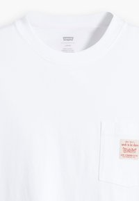White cotton T-shirt with a crew neck, featuring a left chest pocket and a small label with red text. Loose fit design.