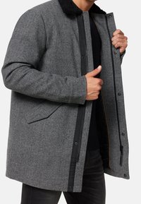 Abrigo de espiga gris con cuello de piel sintética negra. Cuenta con un cierre de cremallera, bolsillos laterales y tejido texturizado. Forro negro visible en el interior.
