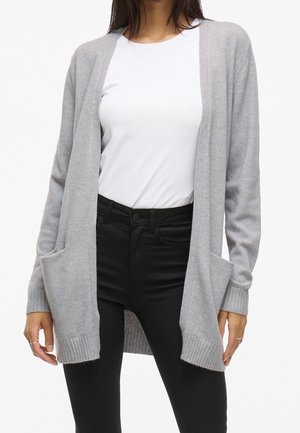 Frau trägt eine hellgraue, offen frontale Strickjacke mit Taschen vorne, ein weißes Oberteil und hoch taillierte schwarze Hose, steht vor weißem Hintergrund.