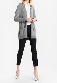 Cardigan lavorato a maglia grigio chiaro con due tasche frontali, design aperto, indossato sopra una camicia bianca a fantasia e pantaloni neri a sigaretta, abbinato a tacchi argento.