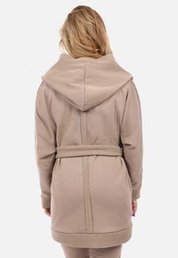 Robe beige con cappuccio dal tessuto morbido, con vita con cintura e cappuccio e polsini foderati in pile a contrasto. Dettaglio della cucitura posteriore visibile.