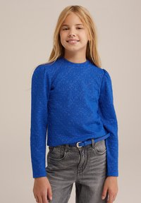 Blå långärmad topp med texturerad blommönster, kompletterad med ett matchande blått bälte. Kombinerad med grå högmidjade jeans.