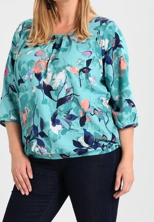 Vrouw draagt prachtige turquoise blouse met een abstract bloemenpatroon in marineblauw, roze en wit, gecombineerd met een donkere spijkerbroek, staand met haar handen ontspannen langs haar zij.