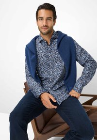Camicia abbottonata con un motivo floreale blu, sovrapposta a un maglione lavorato a maglia blu navy drappeggiato sulle spalle. Jeans blu scuro, seduto su una sedia di legno.