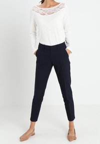 Witte lange mouwen blouse met kantaccent op de halslijn, gecombineerd met marineblauwe tapered broek. Model draagt beige instappers. Effen achtergrond.