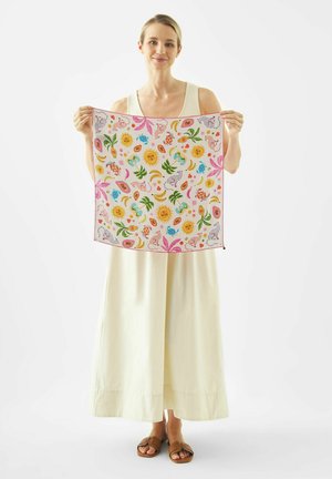 Woman in white dress holding a colorful square scarf with sun, plants, fruits, and animal patterns against a plain background.