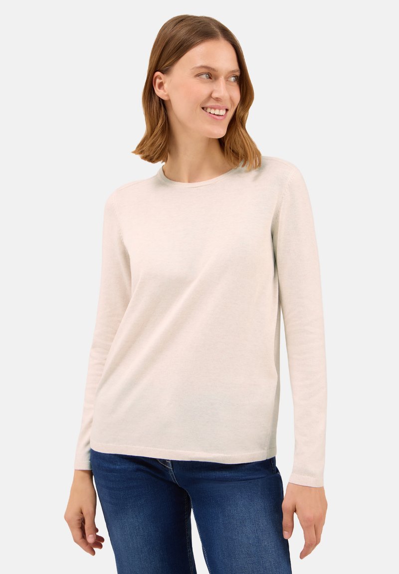 Donna con capelli di lunghezza media che sorride e guarda a destra, indossando un maglione a maniche lunghe beige chiaro e jeans blu scuro.