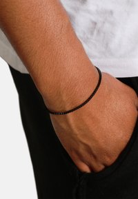 Bracciale di perle nere indossato su un polso, caratterizzato da piccole perle rettangolari lucide con una texture liscia contro la pelle abbronzata.
