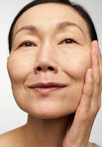 Close-up of a person's face with smooth, tanned skin, light wrinkles, brown eyes, and a subtle smile, hand resting on the cheek.