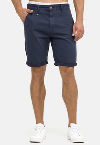Navy-blue cotton shorts with a regular fit, featuring rolled cuffs, a front button closure, and visible side pockets. White sneakers worn below.