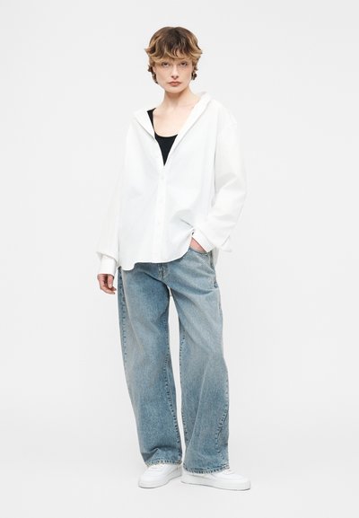 Νεαρό άτομο που φοράει άνετο λευκό πουκάμισο με κουμπιά πάνω από μαύρο τοπ, ανοιχτό μπλε τζιν με φαρδιά μπατζάκια και λευκά αθλητικά παπούτσια, στέκεται μπροστά σε ουδέτερο φόντο.