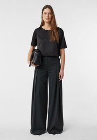 Frau steht da, trägt ein schwarzes kurzärmeliges Oberteil und weite dunkelgraue Hosen, hält eine schwarze Handtasche vor einem schlichten Hintergrund.