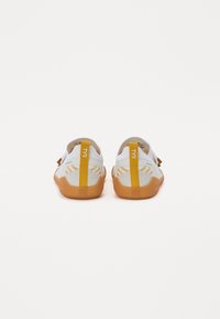 Ζευγάρι άσπρων αθλητικών παπουτσιών με πορτοκαλί σόλες και κίτρινες λαβές, εμφανισμένα από πίσω σε λευκό φόντο.