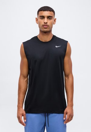 Mladý muž v čiernom tielku Nike bez rukávov a modrých šortkách stojaci tvárou vpred na jednoduchom pozadí.