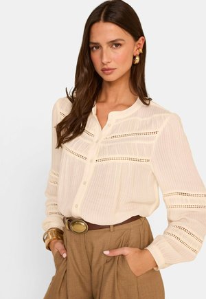Femme aux longs cheveux bruns portant un chemisier crème à boutons avec des détails en dentelle, un pantalon marron taille haute, une ceinture dorée, un bracelet et des boucles d'oreilles.