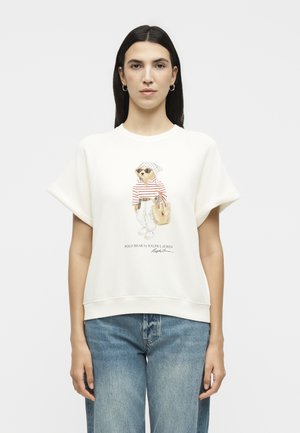 Γυναίκα που φοράει λευκό φούτερ με κοντά μανίκια με γραφικό ενός αρκούδου με γυαλιά ηλίου και ριγέ πουκάμισο, σε συνδυασμό με μπλε τζιν σε απλό φόντο.